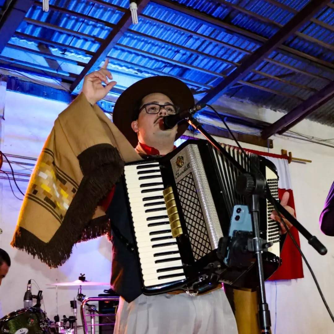 Acordeonista Everton Paschoal, com traje tradicionalista e chapéu, canta e toca acordeona durante uma apresentação musical.