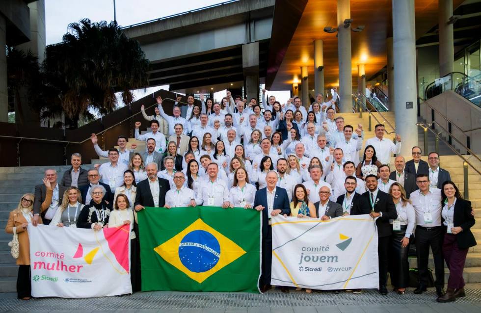 Grupo de representantes do Sicredi posa em escadaria durante evento, segurando a bandeira do Brasil e faixas dos comitês Mulher e Jovem