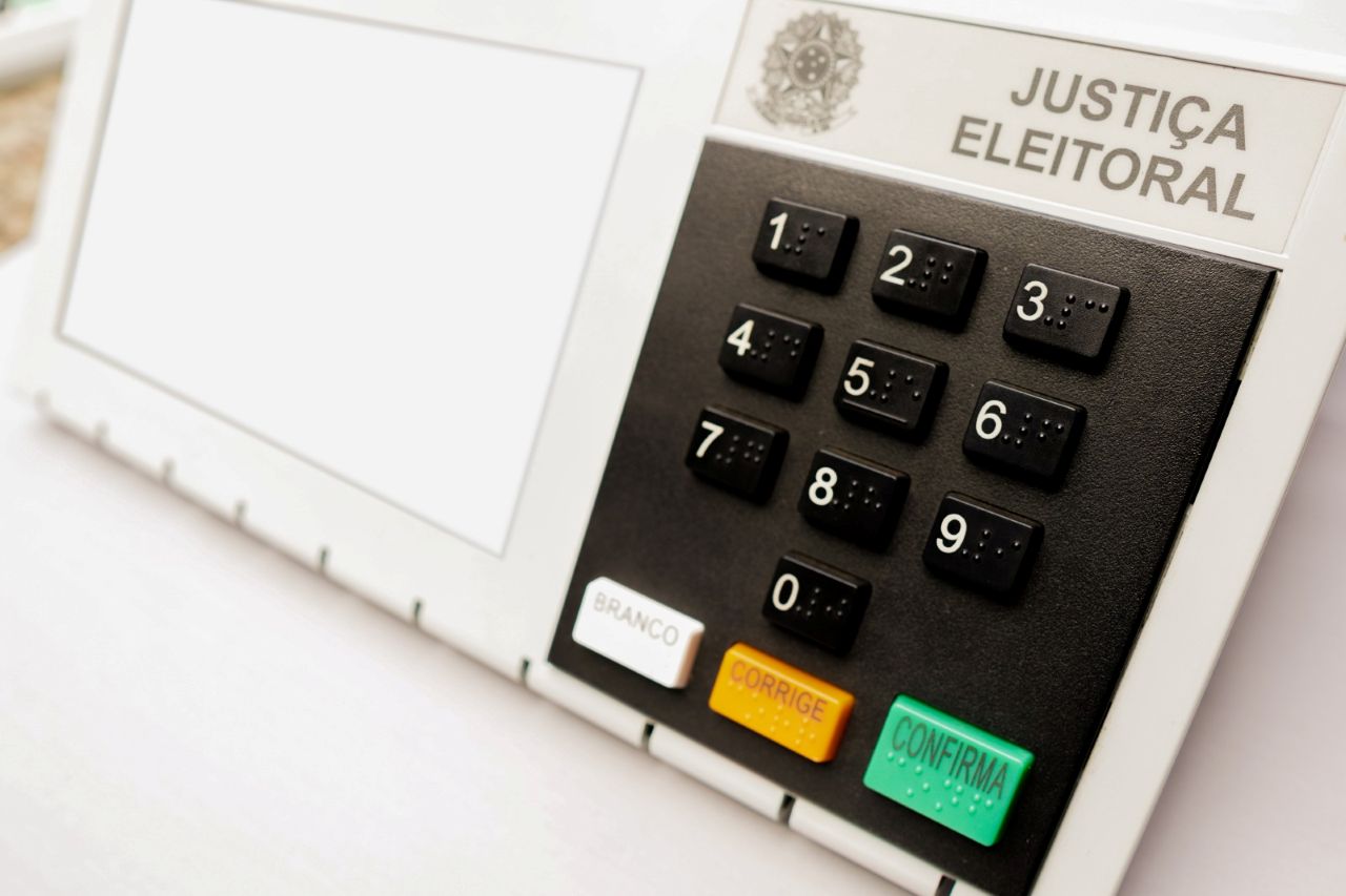 Urna eletrônica da Justiça Eleitoral com tela, teclado numérico em braile e botões branco, corrige e confirma.