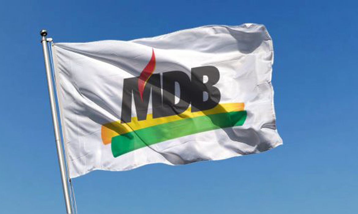 Bandeira branca do MDB com a sigla e as cores verde, amarela e vermelha, hasteada contra o céu azul.