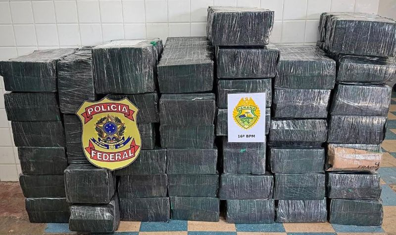 Dezenas de tabletes de maconha embalados e empilhados, com os emblemas da Polícia Federal e do 16º Batalhão da Polícia Militar, após apreensão de 912,1 quilos da droga em Candói.