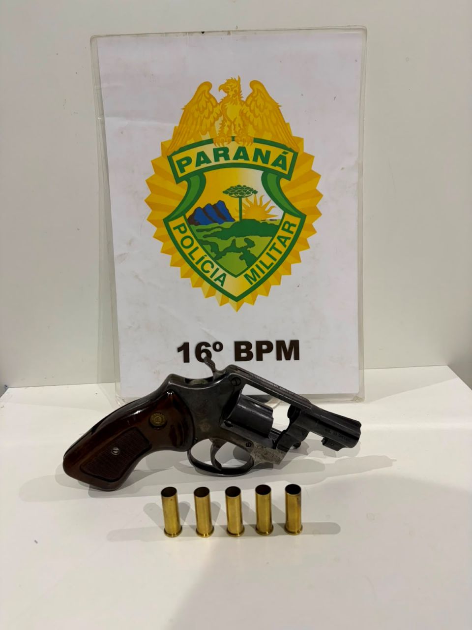 Revólver calibre .38 apreendido pelo 16º BPM, exposto ao lado de cinco estojos de munições deflagradas e diante do brasão da Polícia Militar do Paraná.