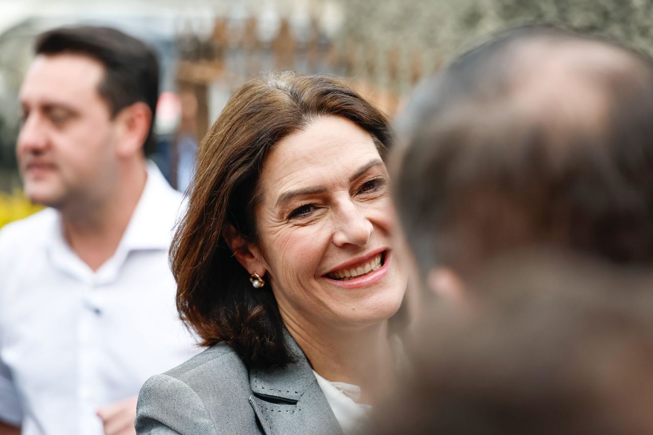 Cristina Graeml, sorridente e usando blazer cinza, conversa com uma pessoa durante um evento ao ar livre.
