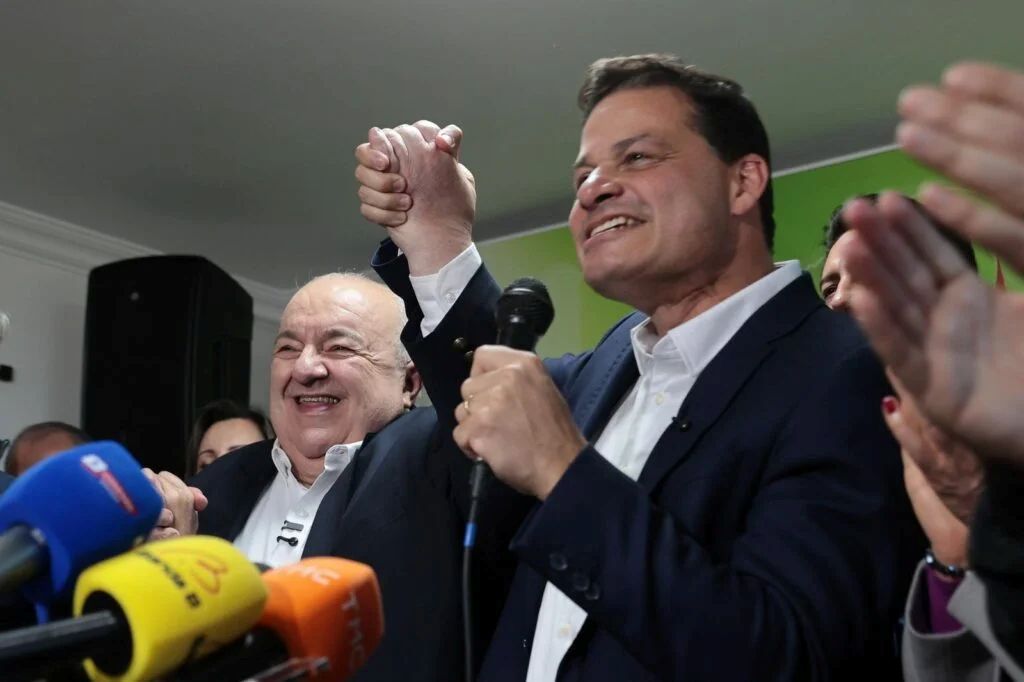 Rafael Greca e Sandro Alex sorriem e levantam as mãos unidas durante evento político, diante de microfones e apoiadores.