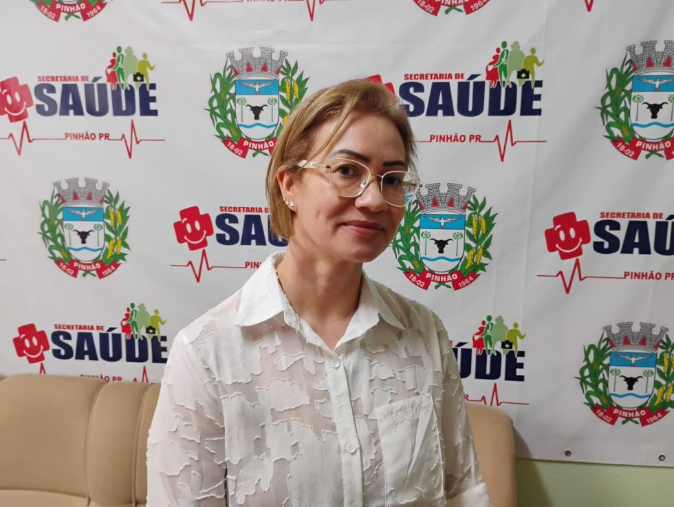 Foto da coordenadora da Atenção Primária da Secretaria Municipal de Saúde de Pinhão, Madalena Zanardini, com cabelos loiros curtos, óculos de grau, vestindo uma camisa branca de manga longa texturizada, sorrindo levemente em frente a um painel institucional da Secretaria de Saúde de Pinhão.