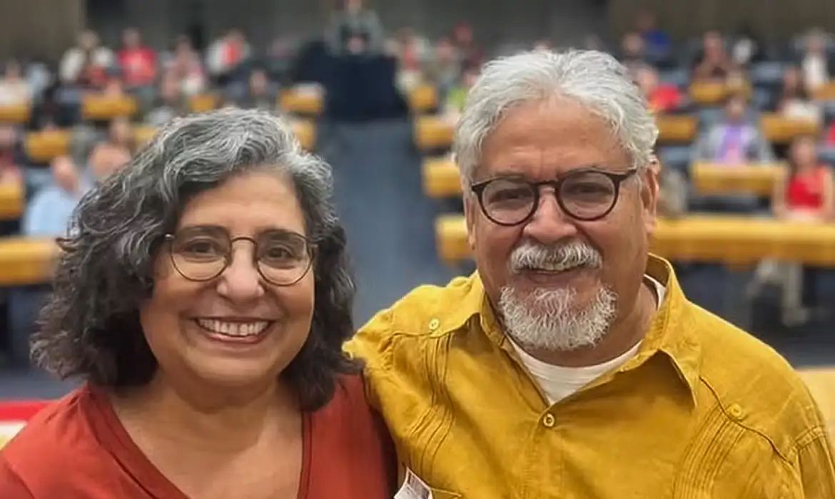 Mulher de blusa vermelha e homem de camisa amarela, ambos de óculos, sorriem lado a lado em um auditório com várias pessoas ao fundo.