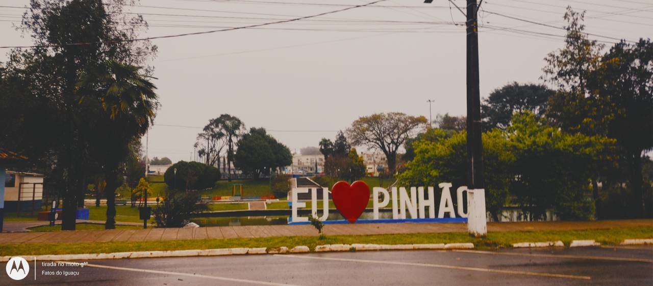 Praça Darci Brolini, em Pinhão, sob céu encoberto, com o letreiro “Eu amo Pinhão” em destaque e a pista molhada pela chuva.