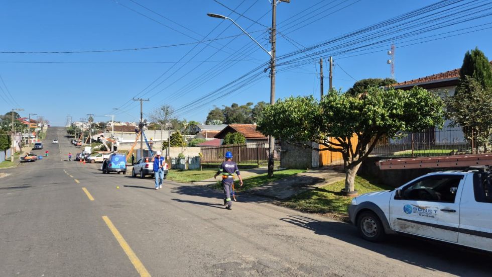 Trabalhadores realizam a regularização de cabos instalados em postes de uma rua residencial de Prudentópolis. Veículos das equipes de serviço estão estacionados e a área de trabalho aparece sinalizada com cones.