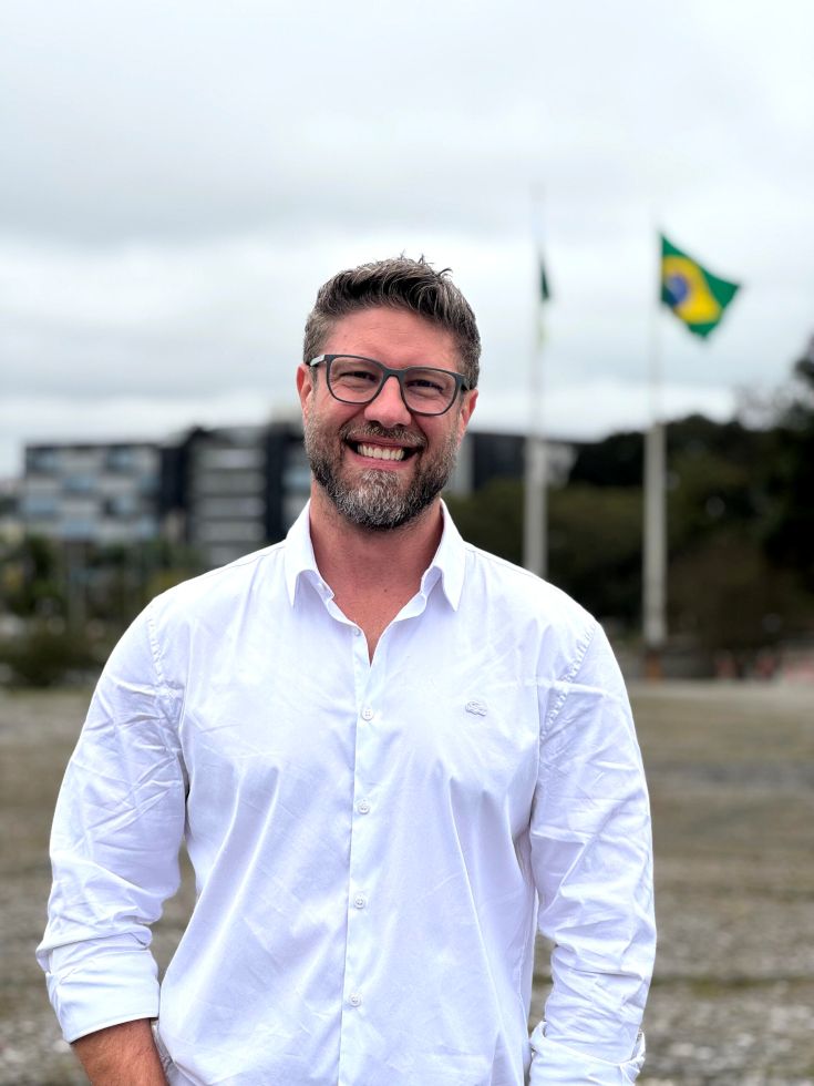 Requião Filho aparece sorrindo, usando óculos e camisa branca, em área externa, com a bandeira do Brasil desfocada ao fundo.