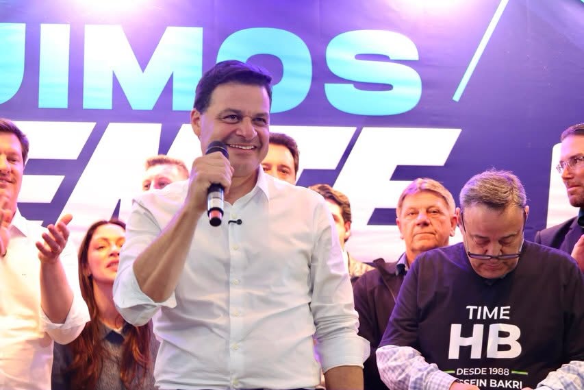 Sandro Alex, sorrindo e segurando um microfone, aparece cercado por apoiadores durante um evento político.