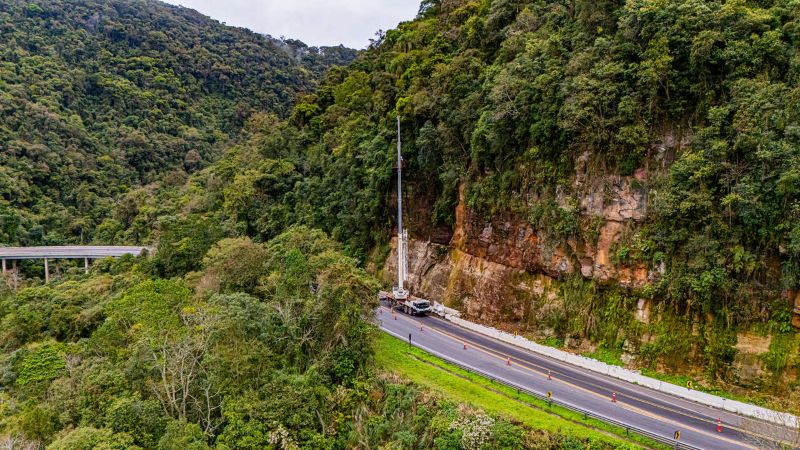 Vista aérea de trecho da BR-277 na Serra da Esperança, cercado por vegetação, com cones sinalizando a pista e equipamento trabalhando junto ao talude.