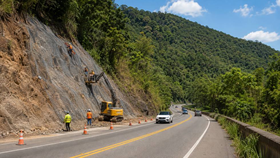 Trabalhadores realizam a estabilização de um talude às margens de uma rodovia cercada por mata, com aplicação de tela de proteção e auxílio de uma escavadeira. Cones sinalizam a área enquanto veículos trafegam pela pista.