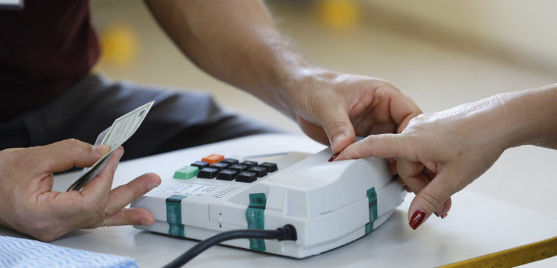 Eleitora pressiona uma tecla da urna eletrônica enquanto outra pessoa segura seu documento de identificação durante o procedimento de votação.
