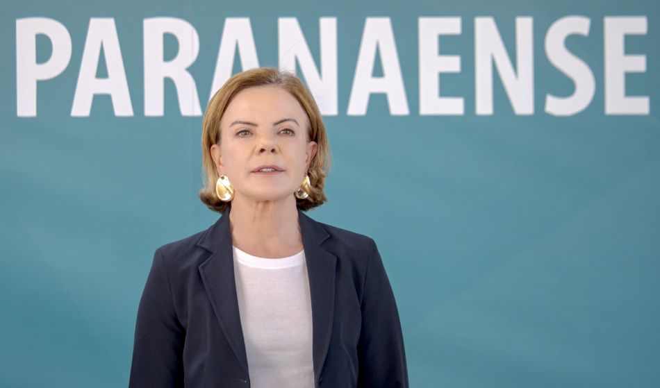 Gleisi Hoffmann aparece diante de um fundo azul, usando camiseta branca e blazer escuro, com a palavra “Paranaense” escrita em destaque.