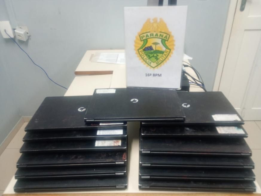 Treze tablets pretos recuperados pela Polícia Militar estão empilhados sobre uma mesa, diante do brasão do 16º BPM.