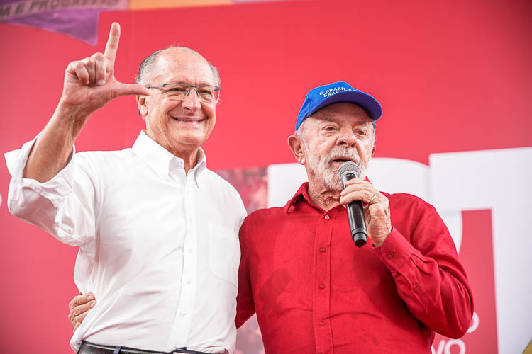 Geraldo Alckmin sorri e faz o “L” com a mão ao lado do presidente Lula, que segura um microfone durante a convenção nacional do PT. Ambos estão diante de um painel vermelho.