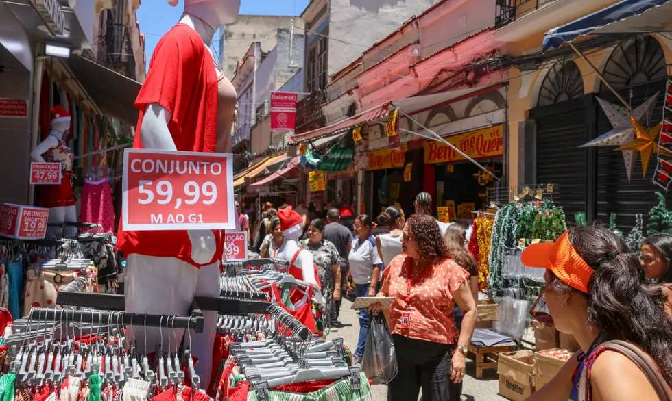 Movimentação intensa de consumidores em uma rua comercial, com lojas de roupas e artigos natalinos expondo produtos e promoções nas calçadas. Manequins, araras e enfeites ocupam os dois lados da via.