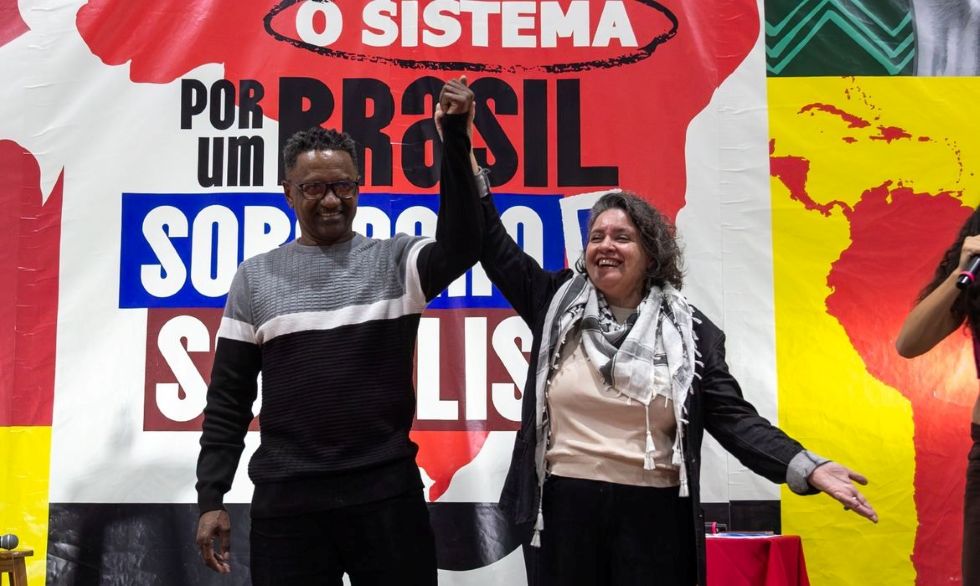 Homem e mulher sorriem e erguem as mãos unidas durante evento político, diante de um painel com a frase “O sistema por um Brasil soberano e socialista”.